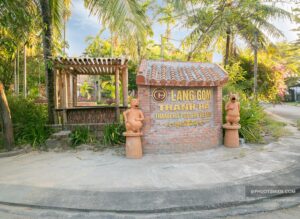 Làng Gốm Thanh Hà với biển hiệu khắc nổi và hai tượng gốm bên lối vào, khung cảnh xanh mát xung quanh.