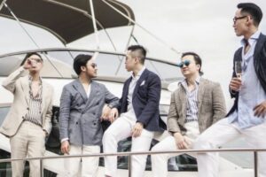 những người đàn ông ăn mặc lịch lãm đứng trên du thuyền, trò chuyện và thưởng thức đồ uống.